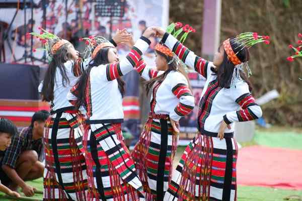 Four young Mizo women with traditional flower-and-feather headpieces are captured performing a dance with interlocking hands, during the 9th State-Level Chapchar Kût 2026, on the outdoor stage at Hmunpui Playground, Jampui Hills, North Tripura.