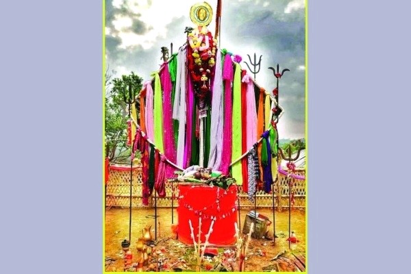 A vibrant Garia Puja deity structure decorated with colorful cloths, garlands, and traditional tridents (Trishul) under a cloudy Tripura sky.