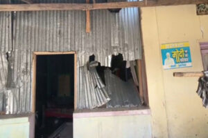 Vandalized house in Tripura after post-poll violence, featuring a damaged corrugated metal wall, broken windows, and a 'Modi Ka Parivar' political poster on an adjacent wall.